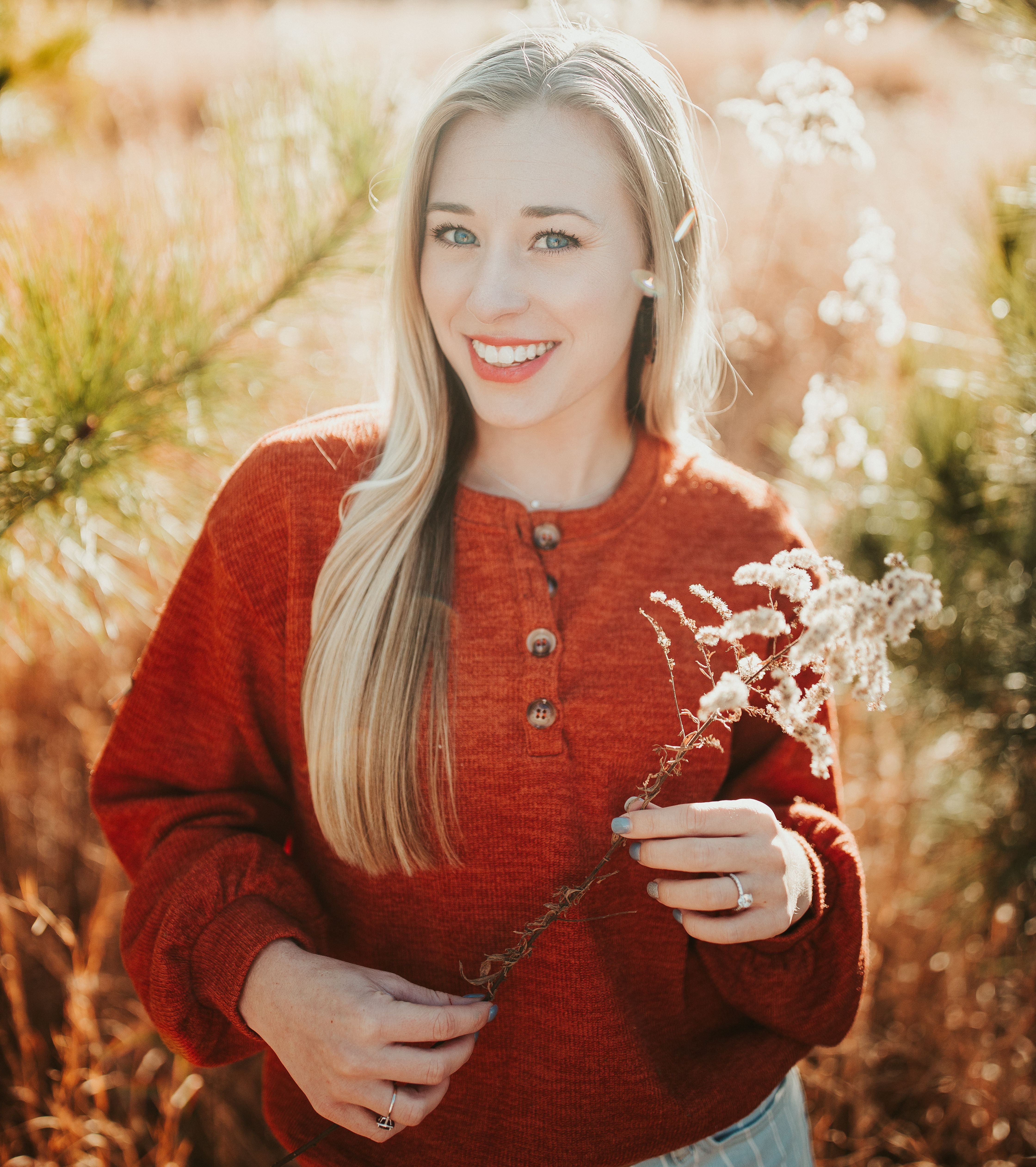 Charming smiling female in red soft sweater standing with plant twig on verdant field on sunny nature and looking at camera happily