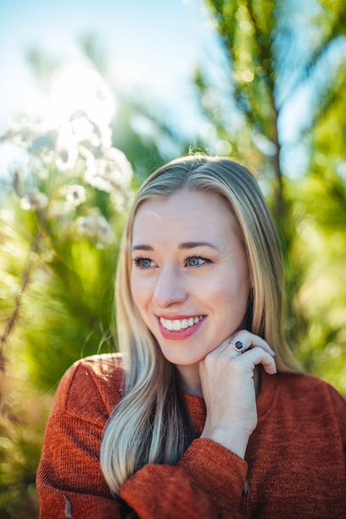 Attractive young blonde in red sweater touching face gently and looking away happily while standing in abundant sunny park