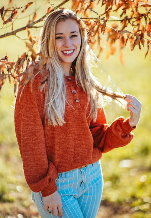 Beautiful cheerful female in warm sweater touching hair and looking at camera with charming smile while standing on sunny autumn park on autumn day