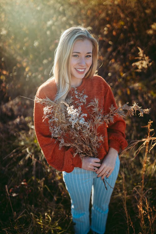 High angle young content female in red sweater standing with dry twigs in hands and looking at camera with toothy smile while standing on grassy sunny field
