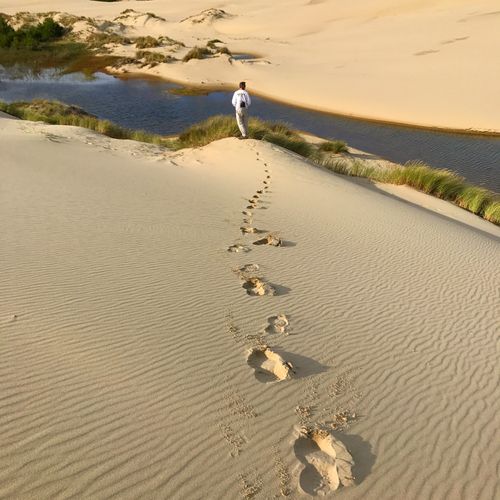 Free A lone figure walking on sand dunes by a lakeside, leaving footprints behind on a sunny day. Stock Photo