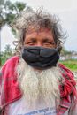 Happy bearded senior Asian male in rustic clothes and protective mask looking at camera while standing on street during COVID pandemic