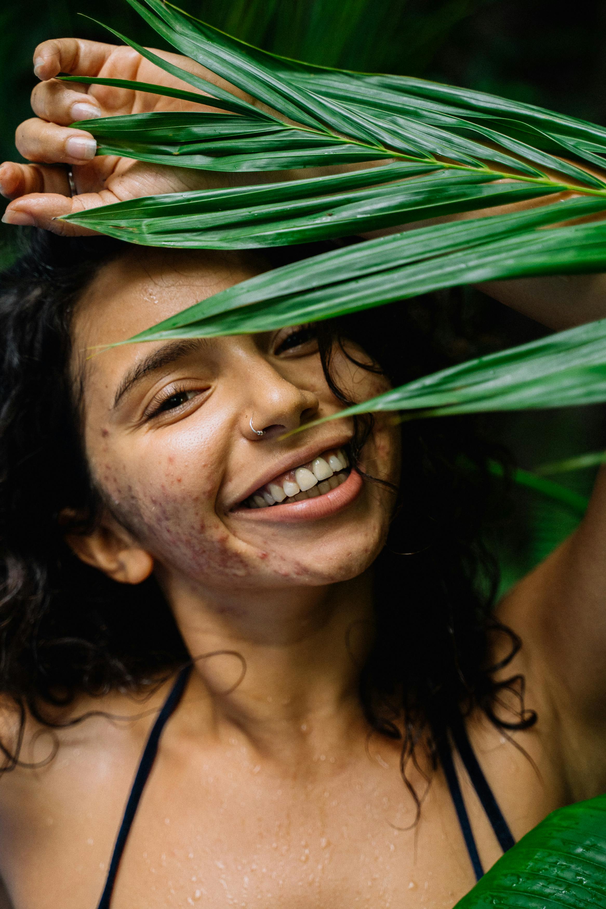 Happy young woman with curly hair and nose piercing surrounded by lush green leaves.
