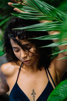 Close-up of woman with curly hair and tattoo posing among vibrant green leaves.