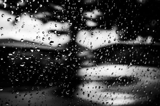Black and white close-up of raindrops on a window with blurred background, creating a moody atmosphere.