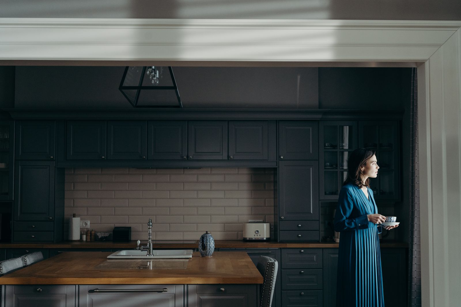 Woman in a long blue dress holding a cup and saucer in a dark kitchen with window light