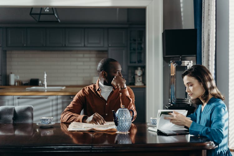 Woman And Man Sitting By Table In Kitchen And Reading Book And Newspaper