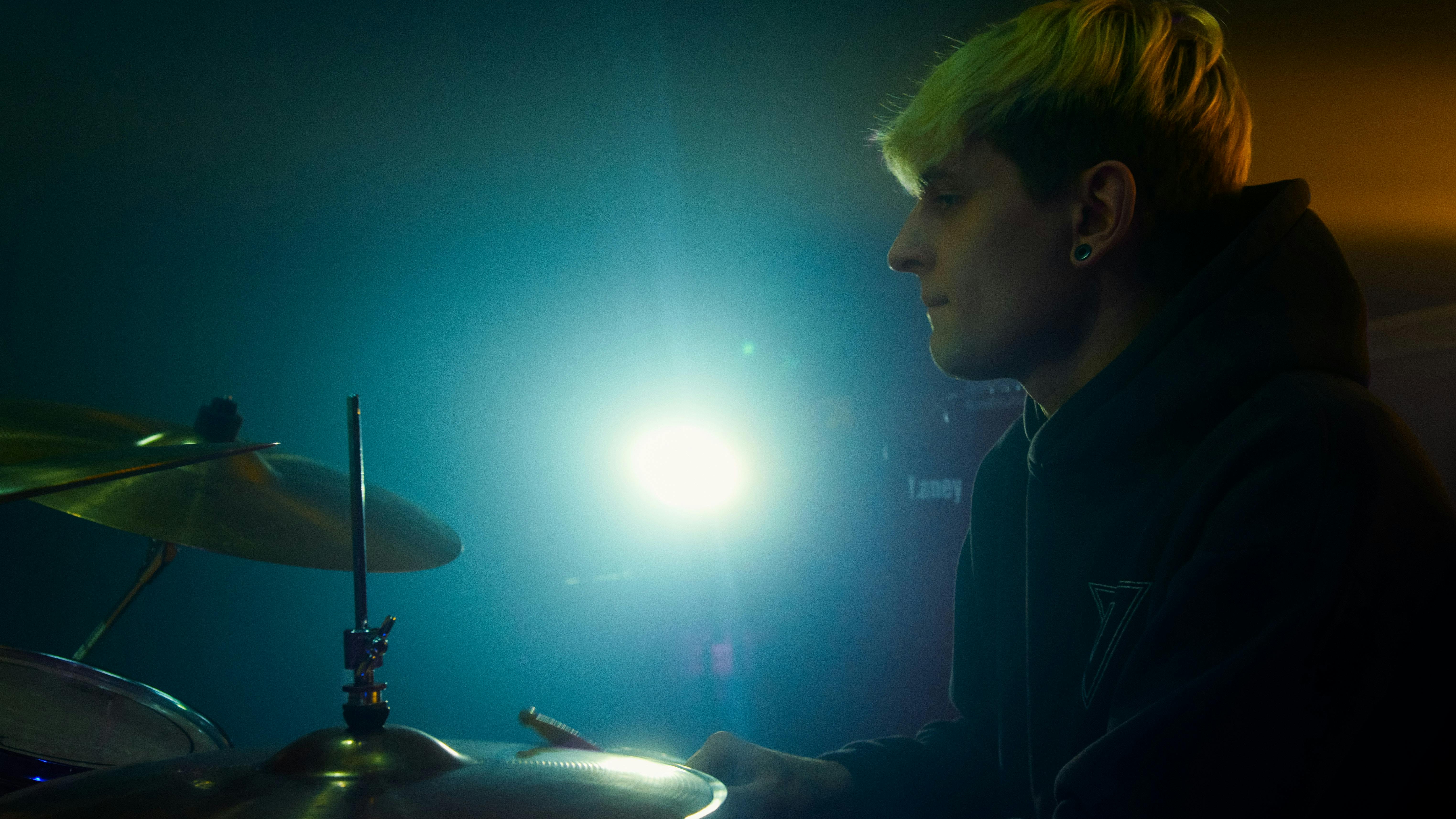 Side view of a young blonde drummer in a hoodie, playing drums under bright stage lights.