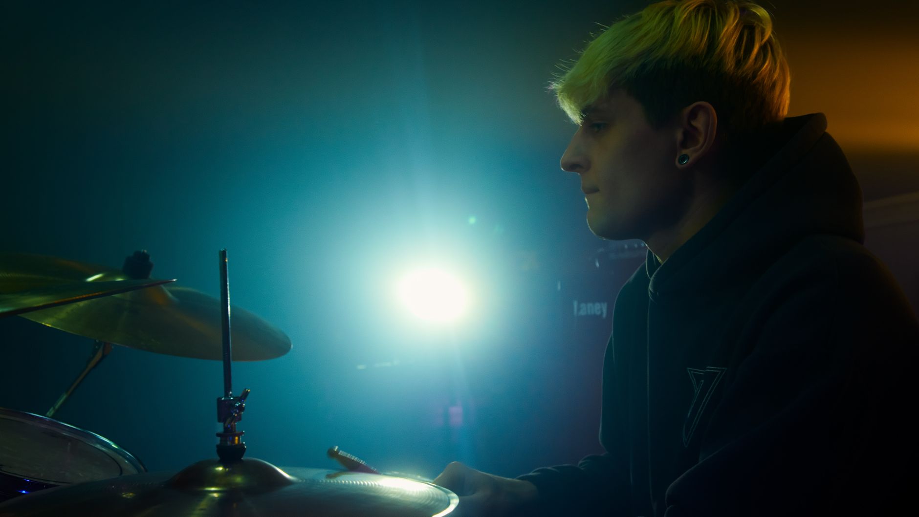演唱會拍攝曝光對焦防閃燈設定指南 Side view of a young blonde drummer in a hoodie, playing drums under bright stage lights.