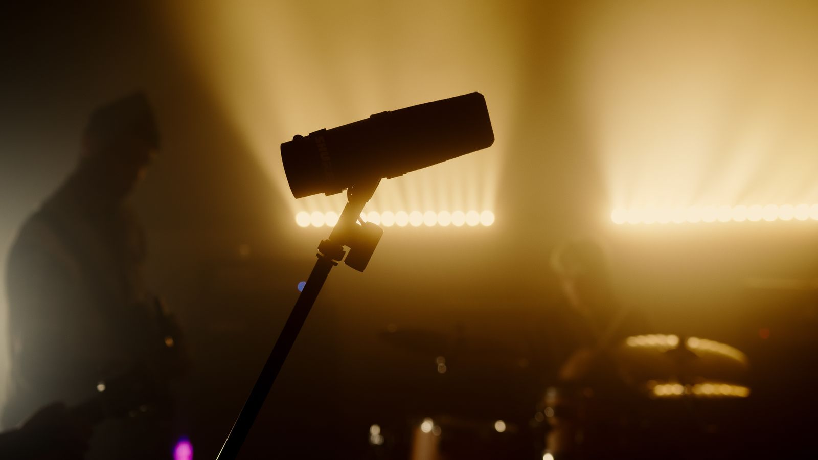 A broadcast microphone on a boom arm in amber stage haze, with a blurred musician behind it