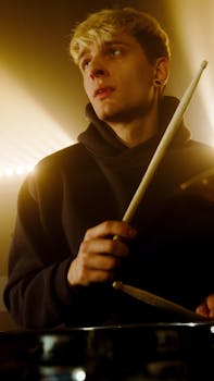 A young adult drummer playing and looking thoughtful under dramatic stage lights.