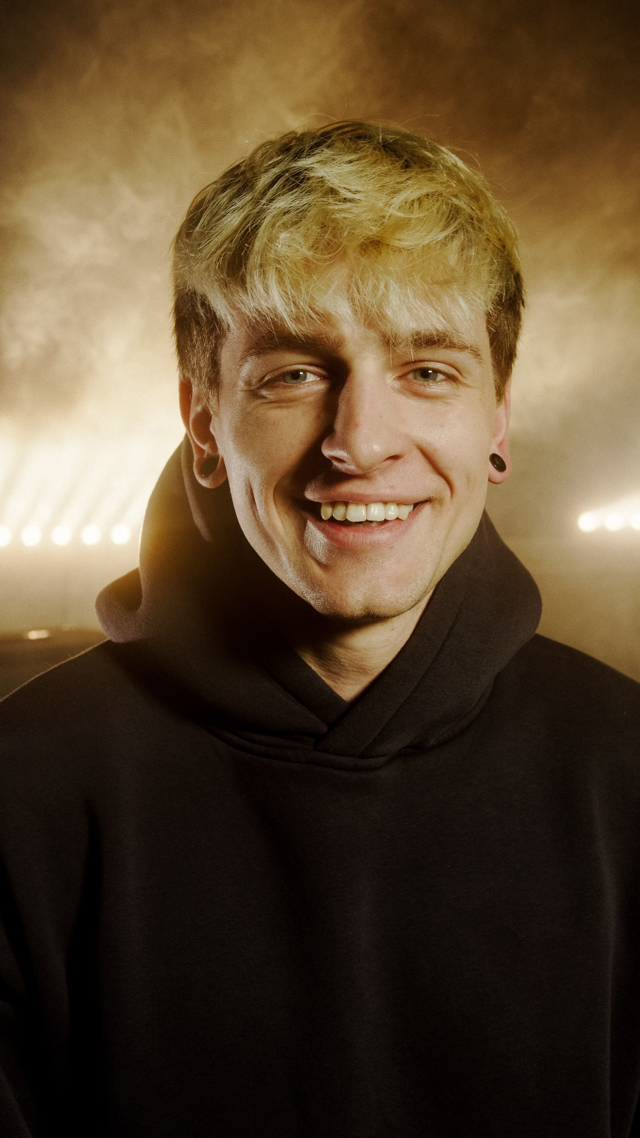 Close-up of a young man smiling in a dark setting with bright lights behind.