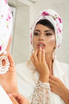 Charming female in turban on head looking at mirror and making makeup in light room in apartment