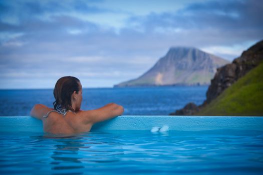Back view of a woman relaxing in an outdoor thermal pool with scenic Icelandic mountains.