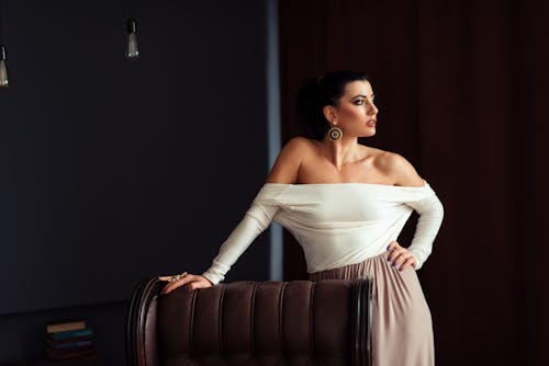 Stylish female with makeup and hand on waist looking away near leather armchair in house