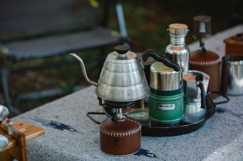 High angle of metal coffee kettle placed on small portable camping gas stove near various utensils on table in nature