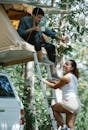 Side view of happy young Hispanic female traveler in casual clothes smiling while climbing ladder towards boyfriend drinking coffee in tent placed on car roof during camping in nature