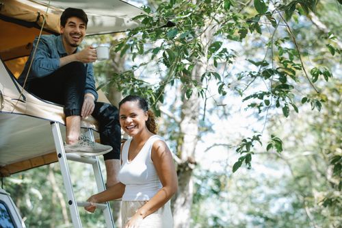 Free Cheerful young Hispanic woman smiling while standing on ladder near positive boyfriend drinking coffee in cam tent during trekking in forest on sunny day Stock Photo