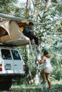 Side view of unrecognizable female camper going upstairs towards boyfriends sitting in tent placed on SUV car roof during road trip in forest on sunny summer day