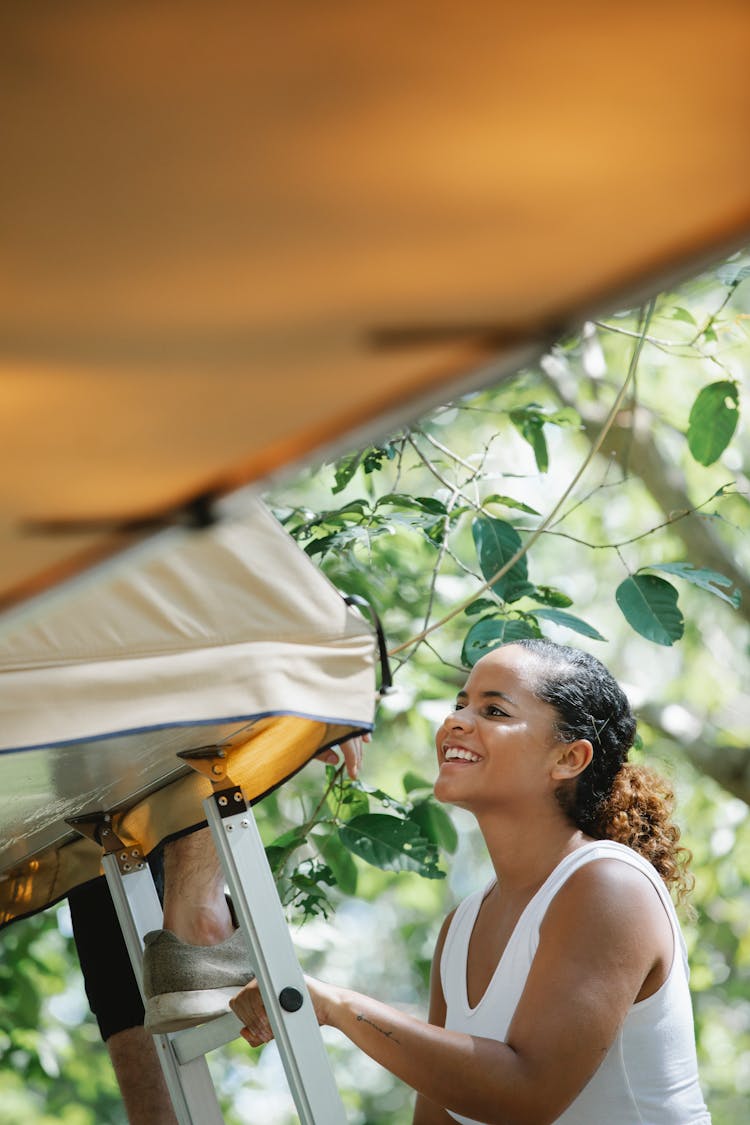 Cheerful Young Hispanic Woman Climbing Ladder Towards Tent During Camping