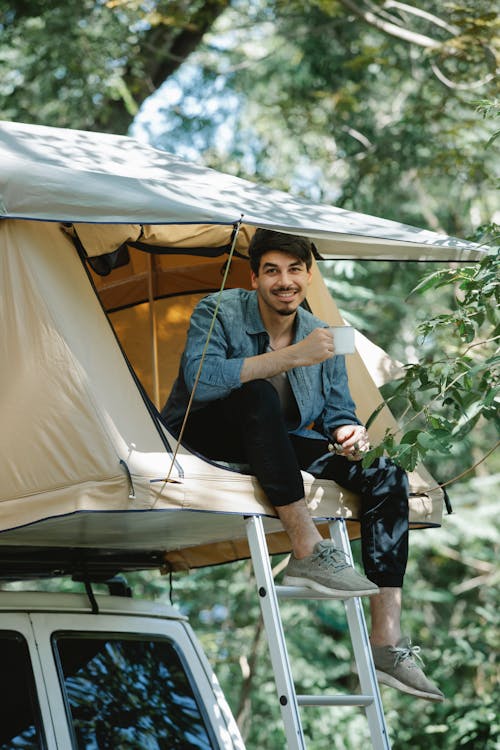 Full body of young delighted young male camper in casual outfit smiling while drinking mug of fresh brewed coffee sitting on tent placed on car roof