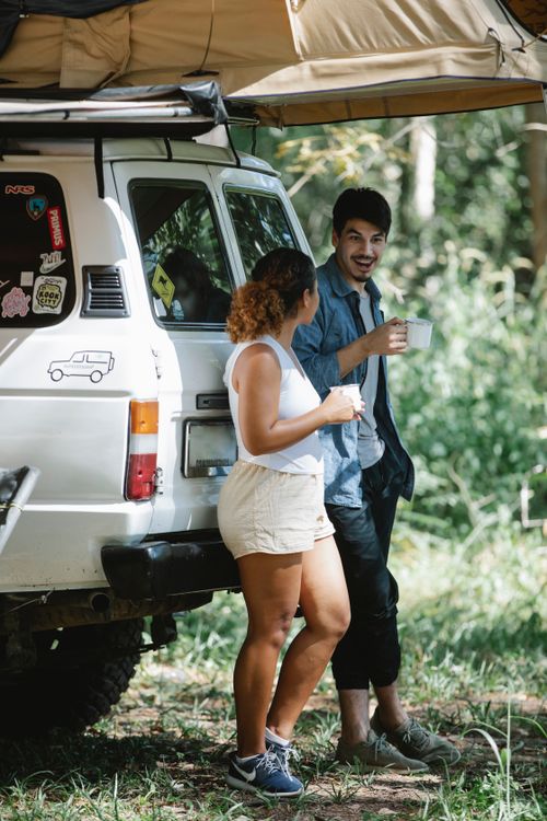 Free Side view of positive romantic young campers in casual clothes leaning on trunk of SUV car and drinking coffee during trip in forest on sunny summer day Stock Photo