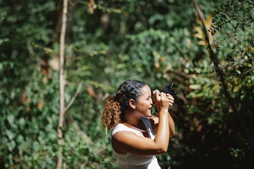 Positive ethnic woman taking photos in lush forest