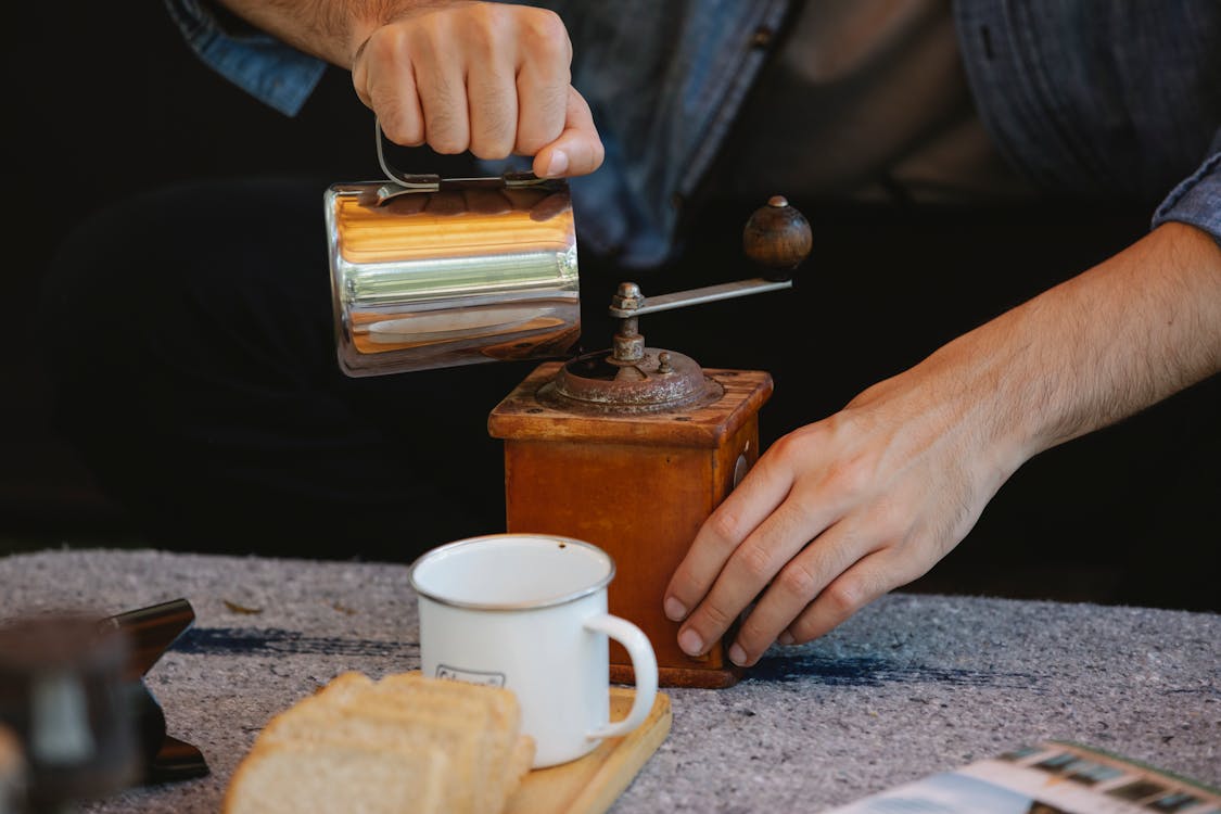 Crop unrecognizable male pouring water into manual coffee grinder placed on table before coffee brewing