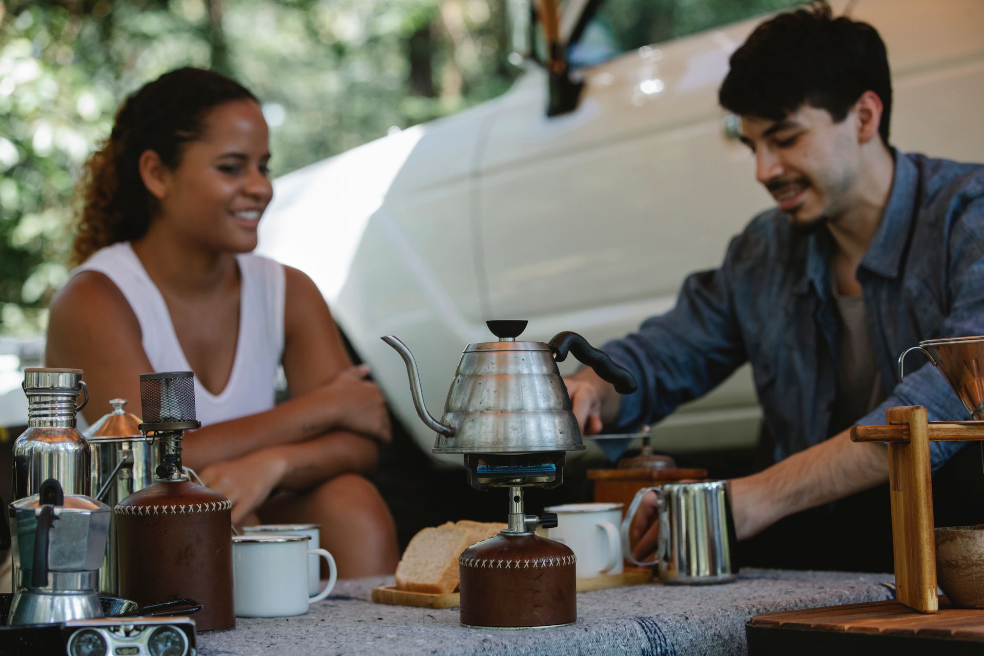 Young cheerful couple in casual wear boiling water in kettle on gas stove and grinding beans for coffee brewing while spending sunny day in camp