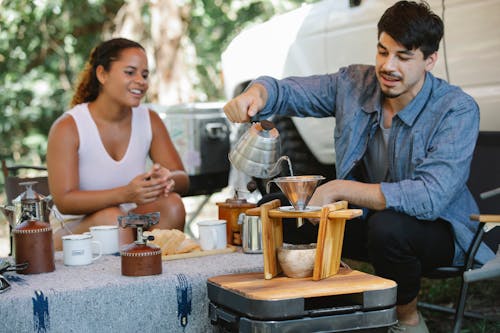 Content young man pouring hot water from gooseneck kettle into filter while preparing pour over coffee with smiling girlfriend during picnic