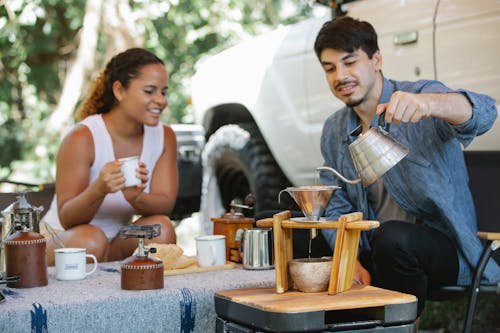Cheerful young multiracial couple in casual clothes pouring boiling hot water into pour over filter while having coffee break together in sunny nature