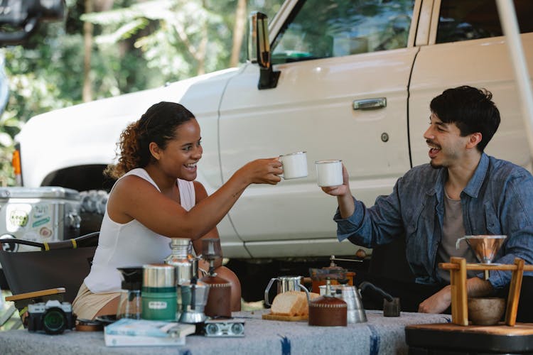 Joyful Young Diverse Couple Clinking Coffee Cups During Picnic In Nature