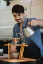 Happy young ethnic male tourist pouring water into cup through metal funnel while brewing coffee during picnic on sunny day