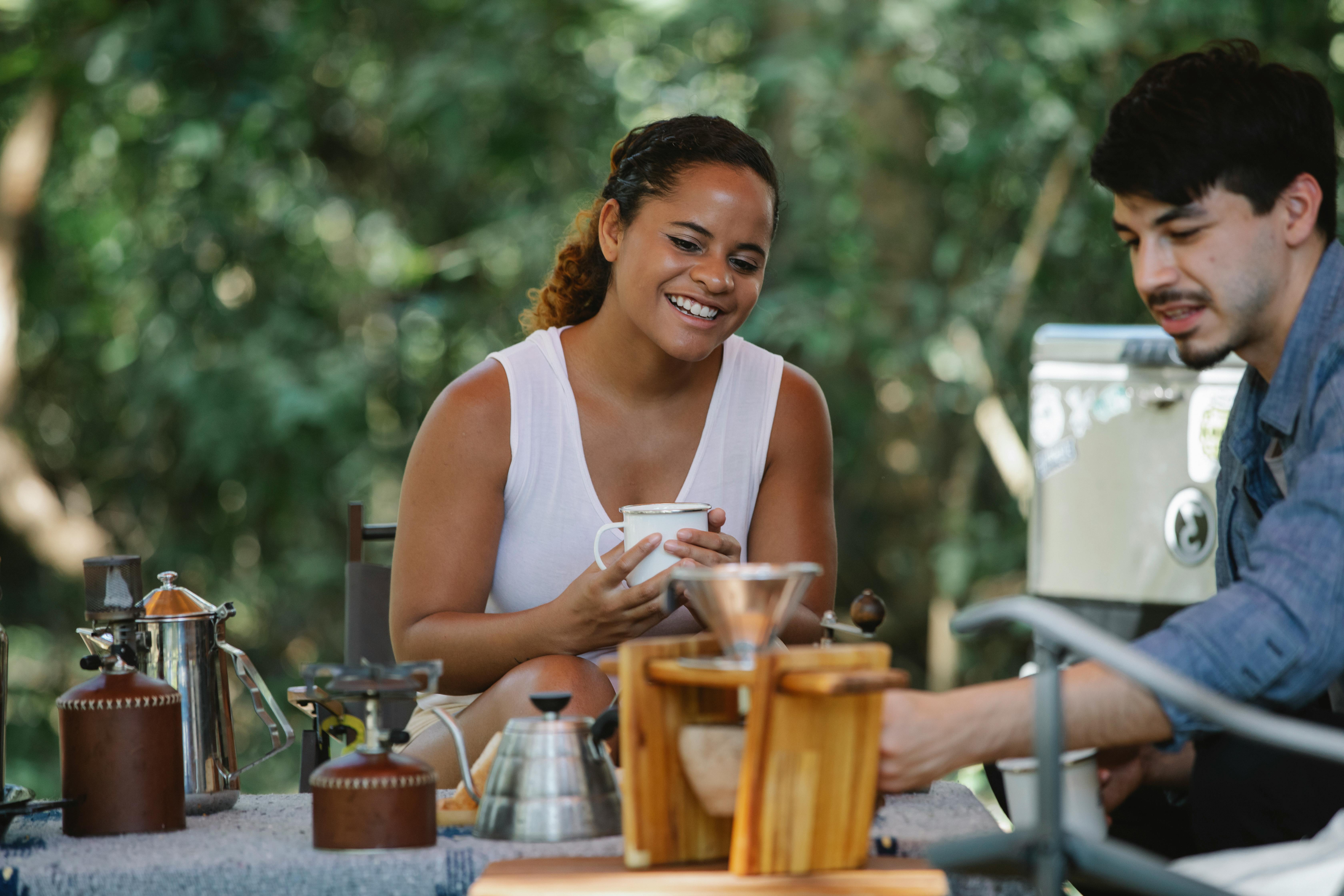 A cheerful couple enjoys freshly brewed coffee at an outdoor campsite, surrounded by nature.