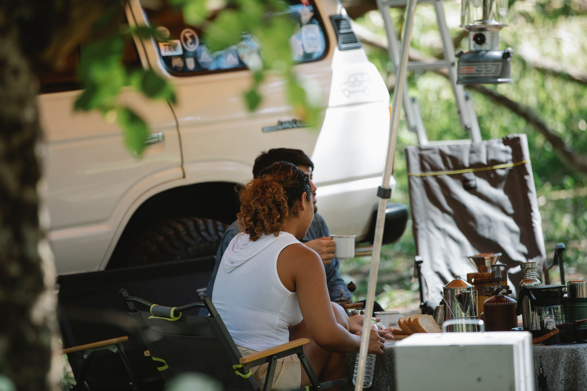 Side view of unrecognizable couple sitting at table near SUV car and drinking coffee during road trip in nature on sunny summer day