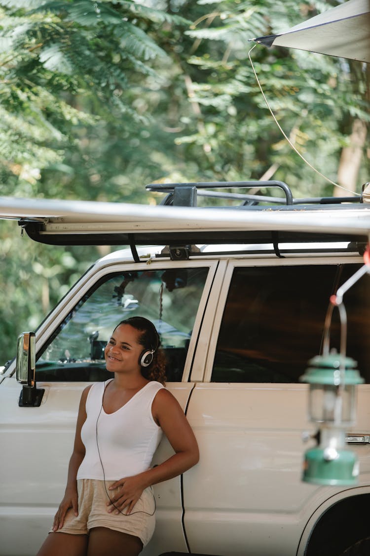 Joyful Ethnic Woman Enjoying Music In Headphones While Leaning On Car At Campsite