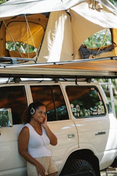 Positive young ethnic lady in casual clothes smiling and leaning on off road car with tent while listening to music in headphone during camping in forest
