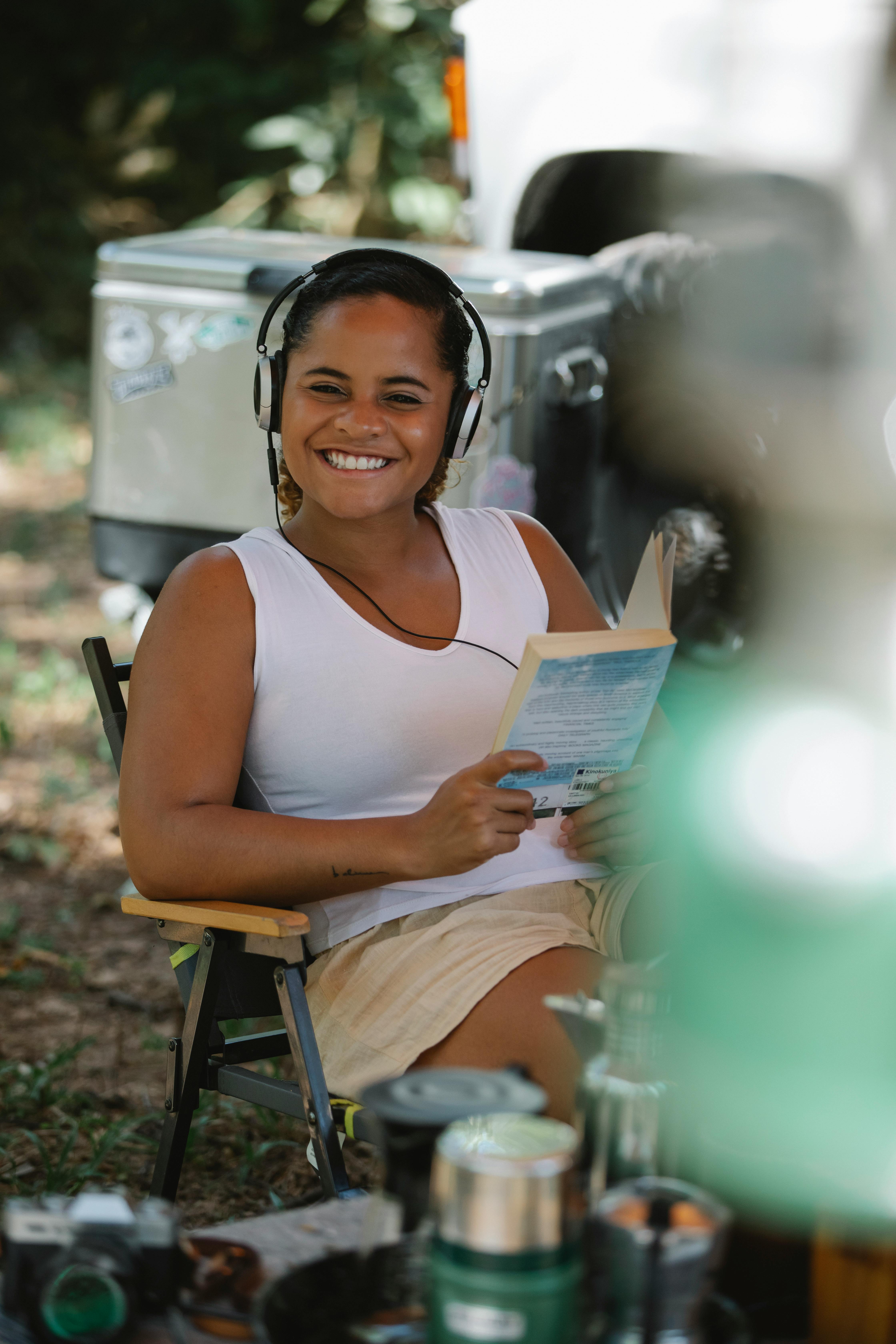 content smiling ethnic lady reading book and listening to music in forest