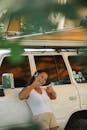 Excited young ethnic female camper showing thumbs up and listening to music in headphones while leaning on car under tent during summer holidays