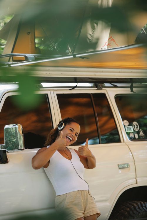 Free Excited young ethnic female camper showing thumbs up and listening to music in headphones while leaning on car under tent during summer holidays Stock Photo