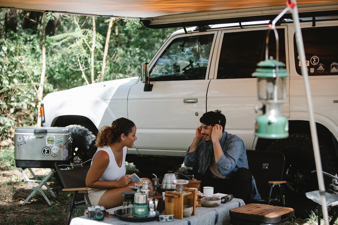 Positive multiethnic friends sitting at table near van with awning in forest
