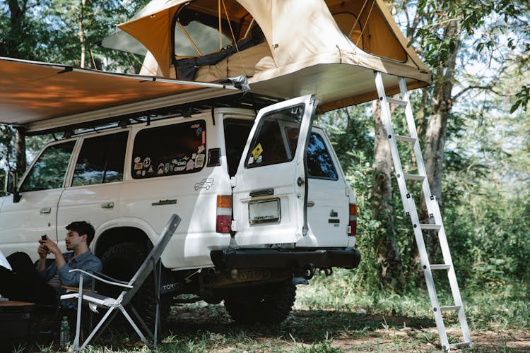 Man Sitting Under Awning Placed Near Offroader With Tent On Roof In Forest