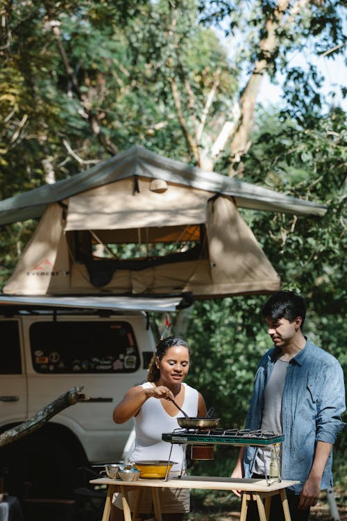 Focused multiracial couple cooking on metal stove using skillet in camp near car and tent