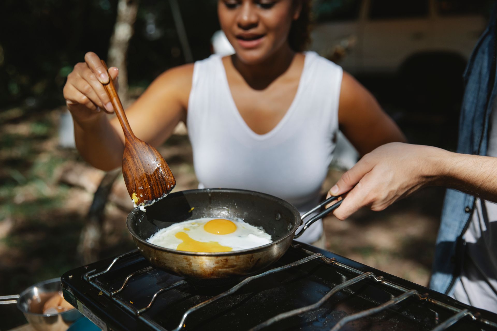 A diverse couple cooking fried eggs together on a portable stove in a forest setting.