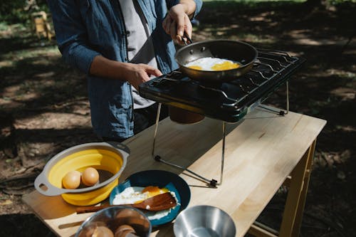 Free From above of crop anonymous male cooker frying eggs on skillet using metal stove on table in forest Stock Photo