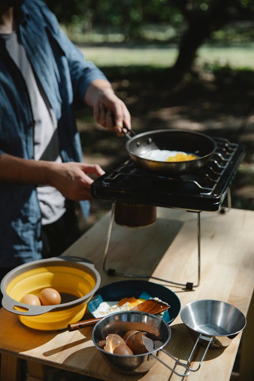 Free High angle of crop unrecognizable male cooker frying eggs on metal burner using skillet in forest Stock Photo