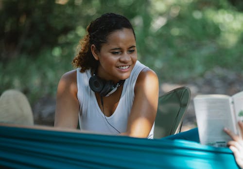 Happy young Hispanic female with headphones sitting on chair near friend in hammock with book while spending time in woods