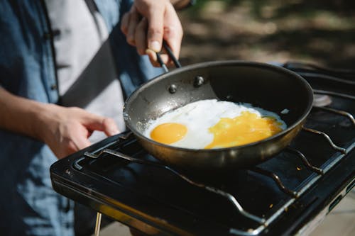 From above of crop faceless male cooker frying eggs on metas gas burner using skillet in nature