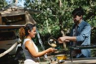 Side view of cheerful young multiracial couple in casual clothes putting scrambled eggs from skillet on plate standing near metal stove in camp