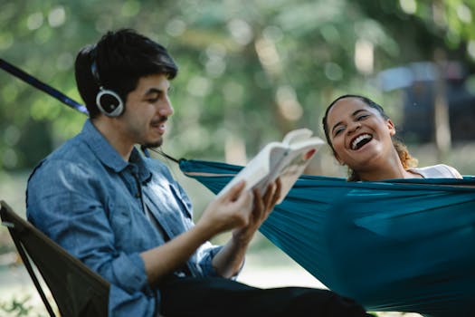 Cheerful young multiracial couple having conversation while resting in forest with book and hammock